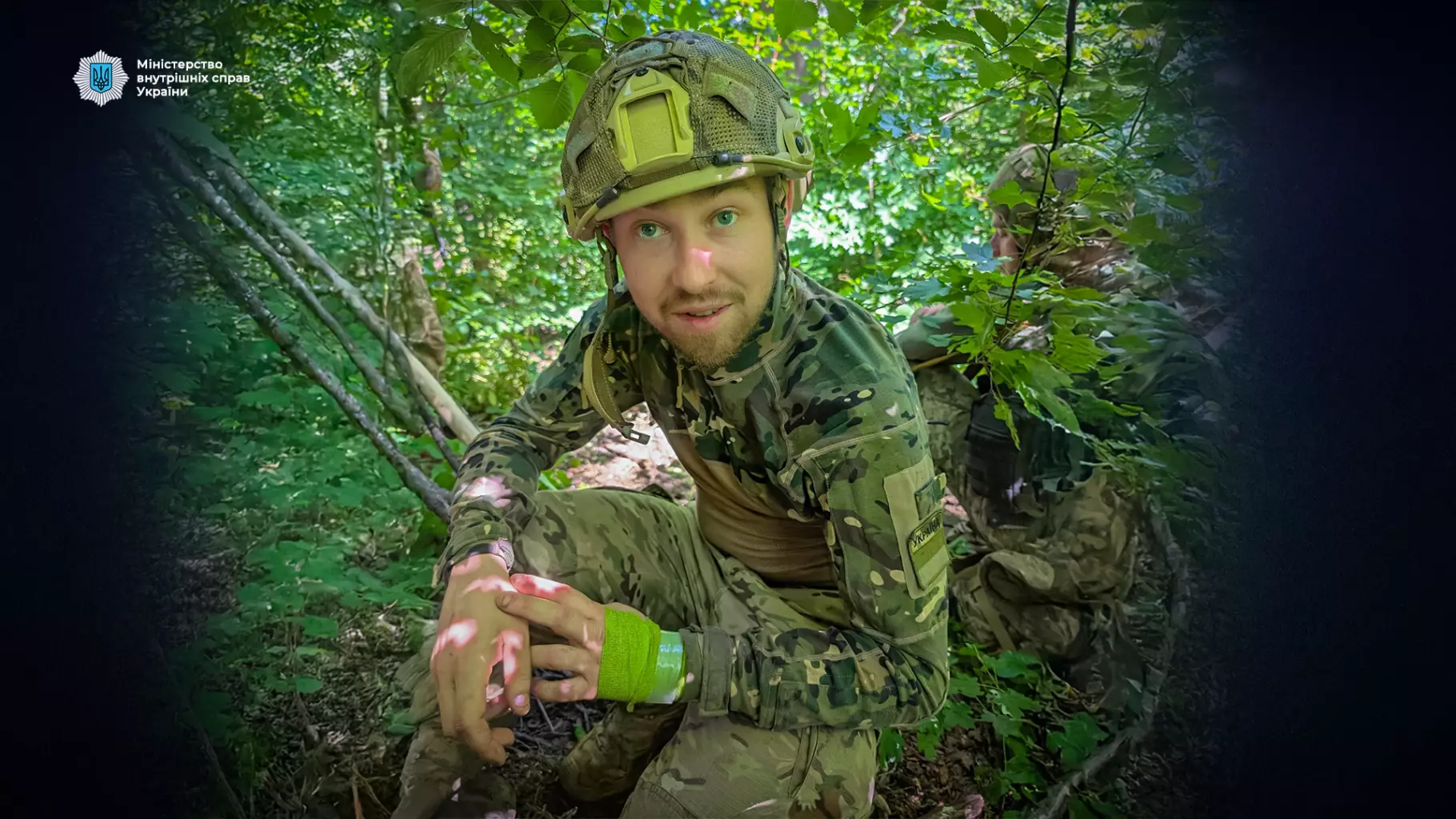 Український прикордонник у камуфляжі сидить у лісі, тримаючи руки на колінах, на тлі зеленого листя