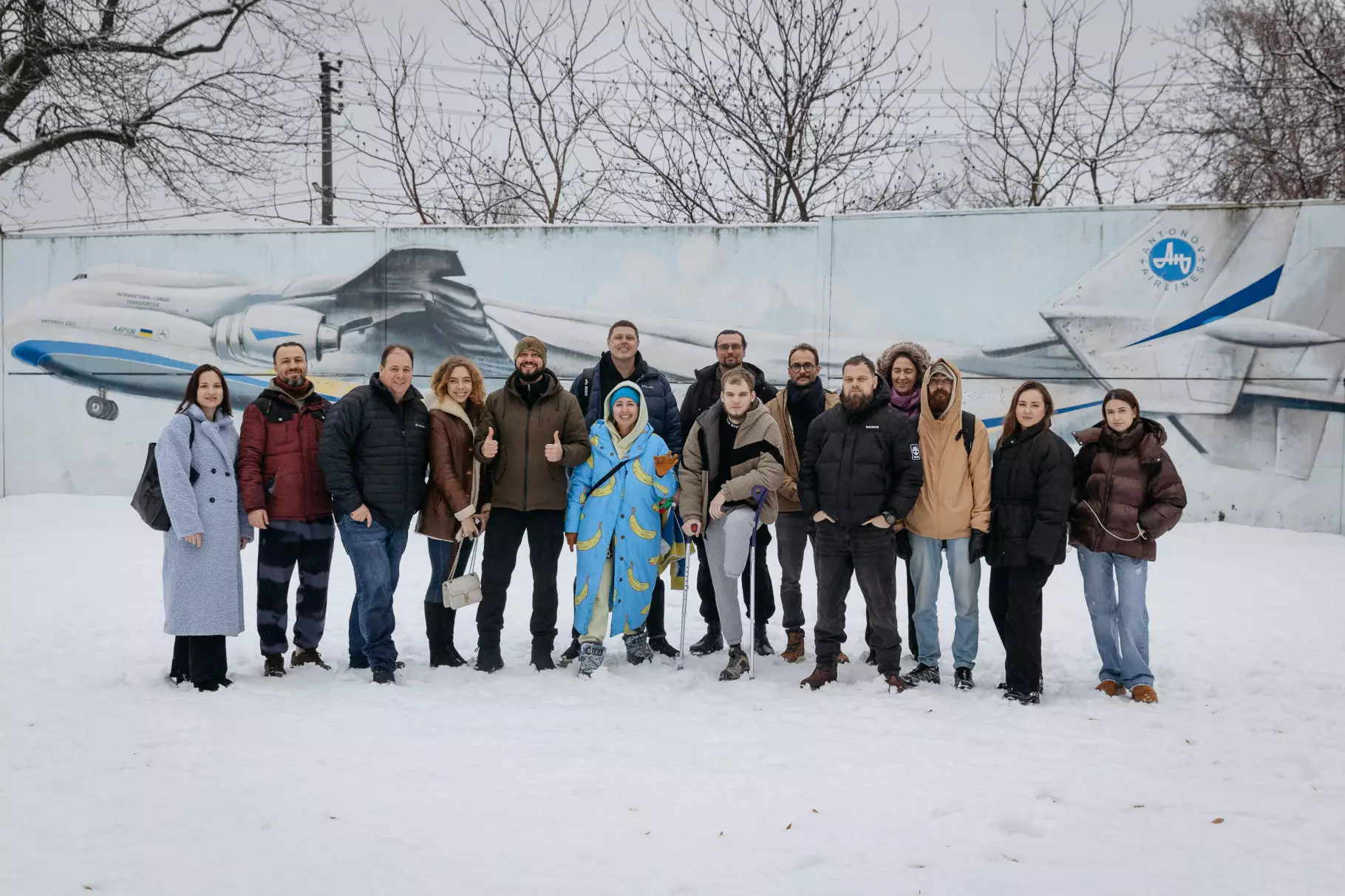 Група людей стоїть на снігу перед стіною з малюнком літака, усі одягнені в зимовий одяг