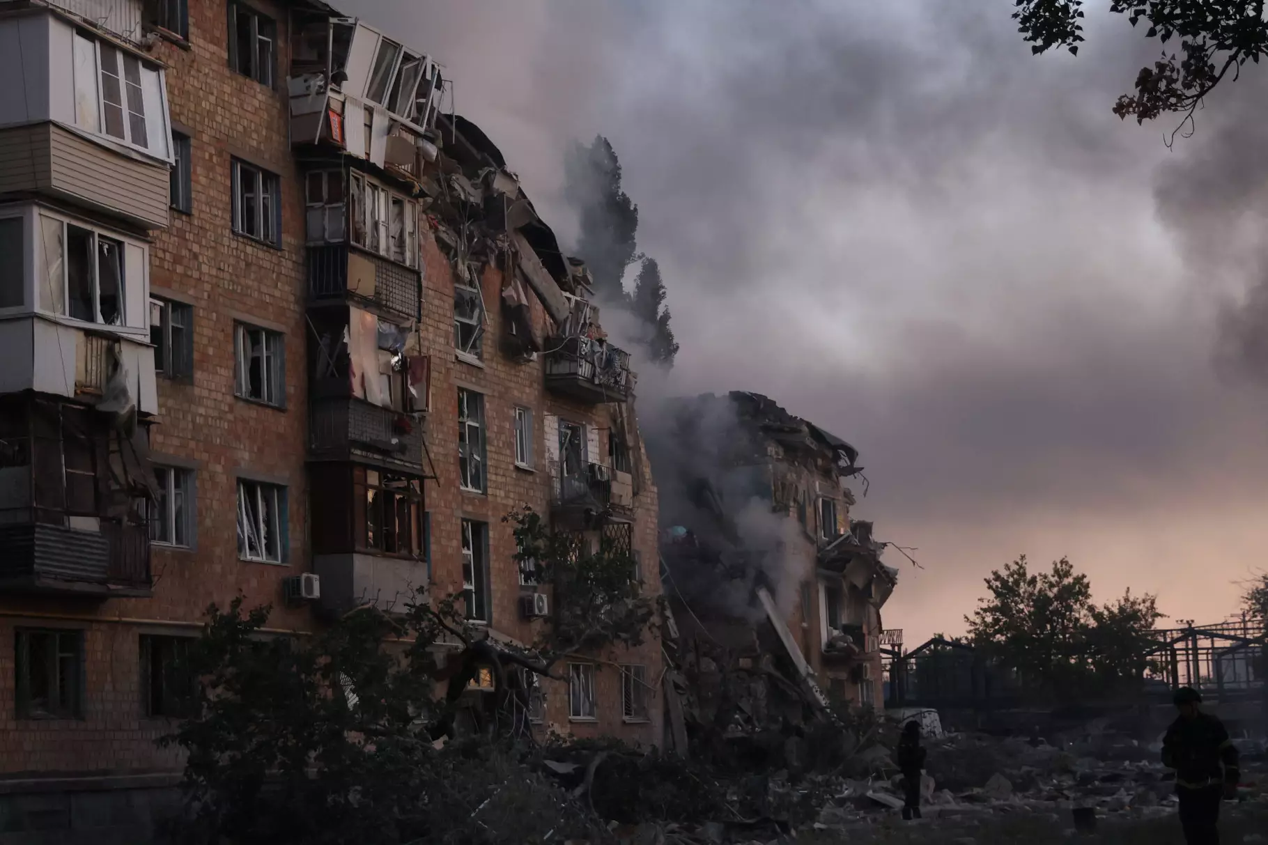 Зруйнована багатоповерхівка після обстрілу, дим піднімається в небо, люди оглядають пошкодження