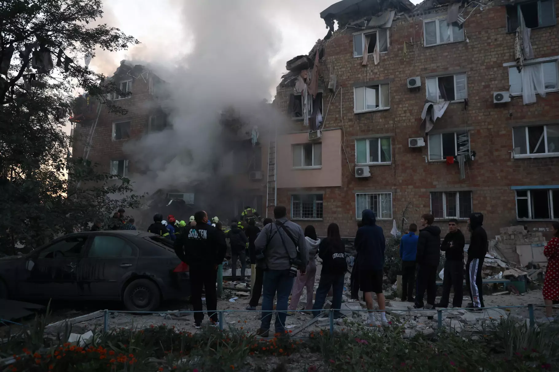 Люди спостерігають за рятувальниками біля пошкодженого обстрілом житлового будинку, дим піднімається вгору
