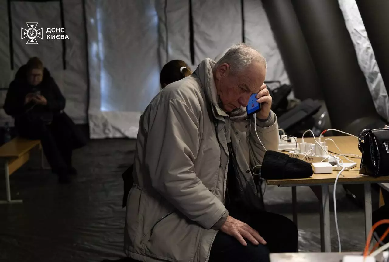 Літній чоловік у пункті обігріву заряджає телефон, сидячи за столом у теплому наметі