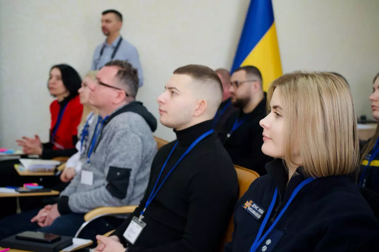 Учасники тренінгу УП+ у МВС України слухають лекцію, на задньому плані прапор України