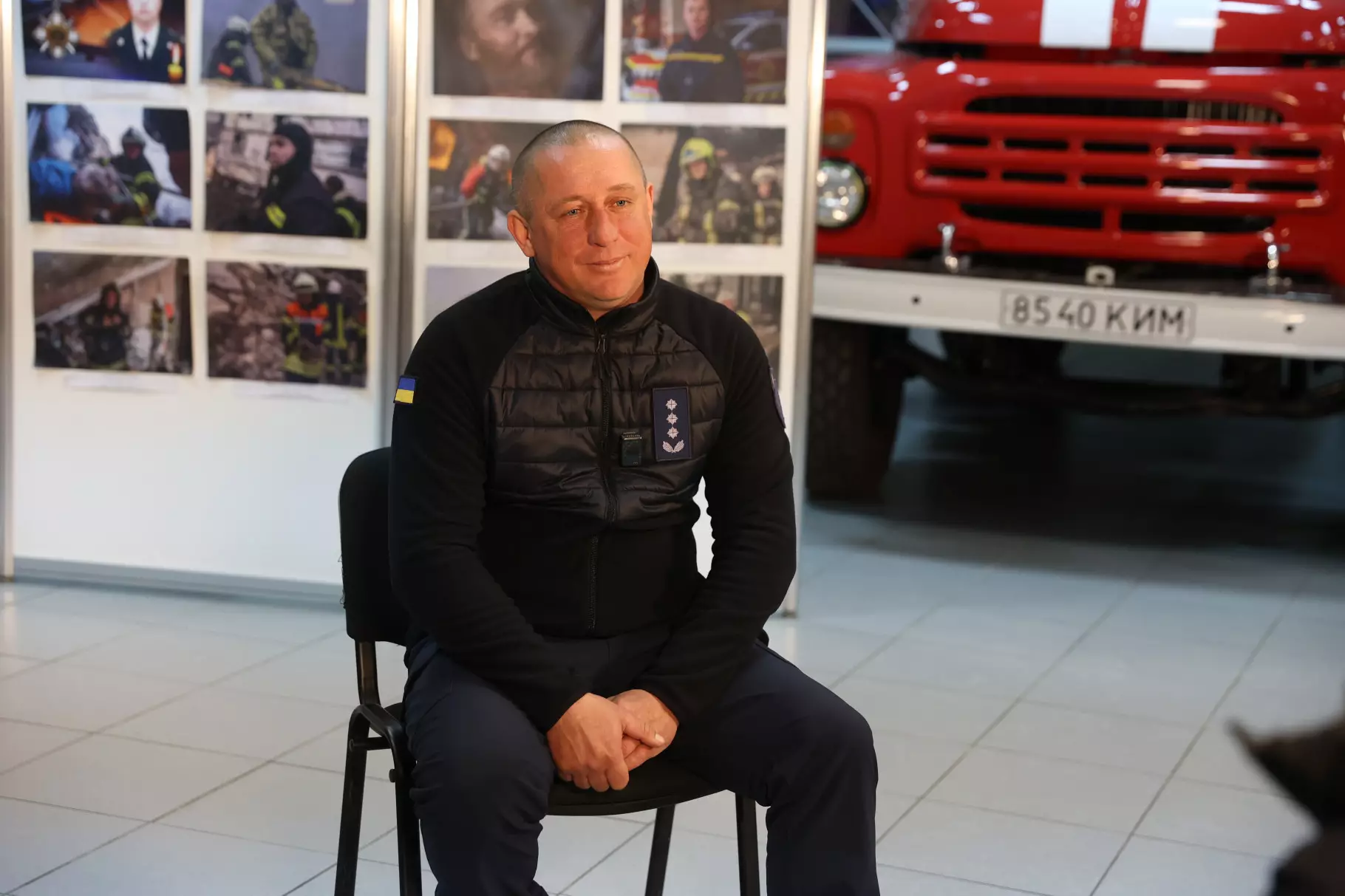 Рятувальник ДСНС України сидить на стільці в приміщенні, на задньому плані червона пожежна машина