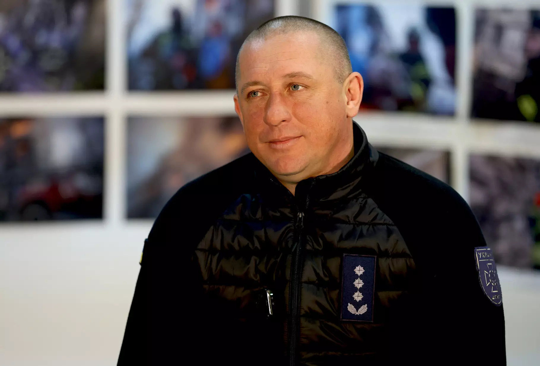 Чоловік у чорній куртці з емблемою ДСНС на фоні фотографій, пов'язаних з рятувальними операціями