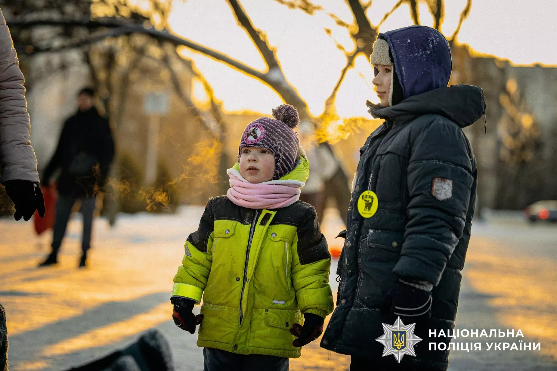 Двоє дітей у зимовому одязі на вулиці, один з них у світловідбивальному жилеті, поруч поліцейський