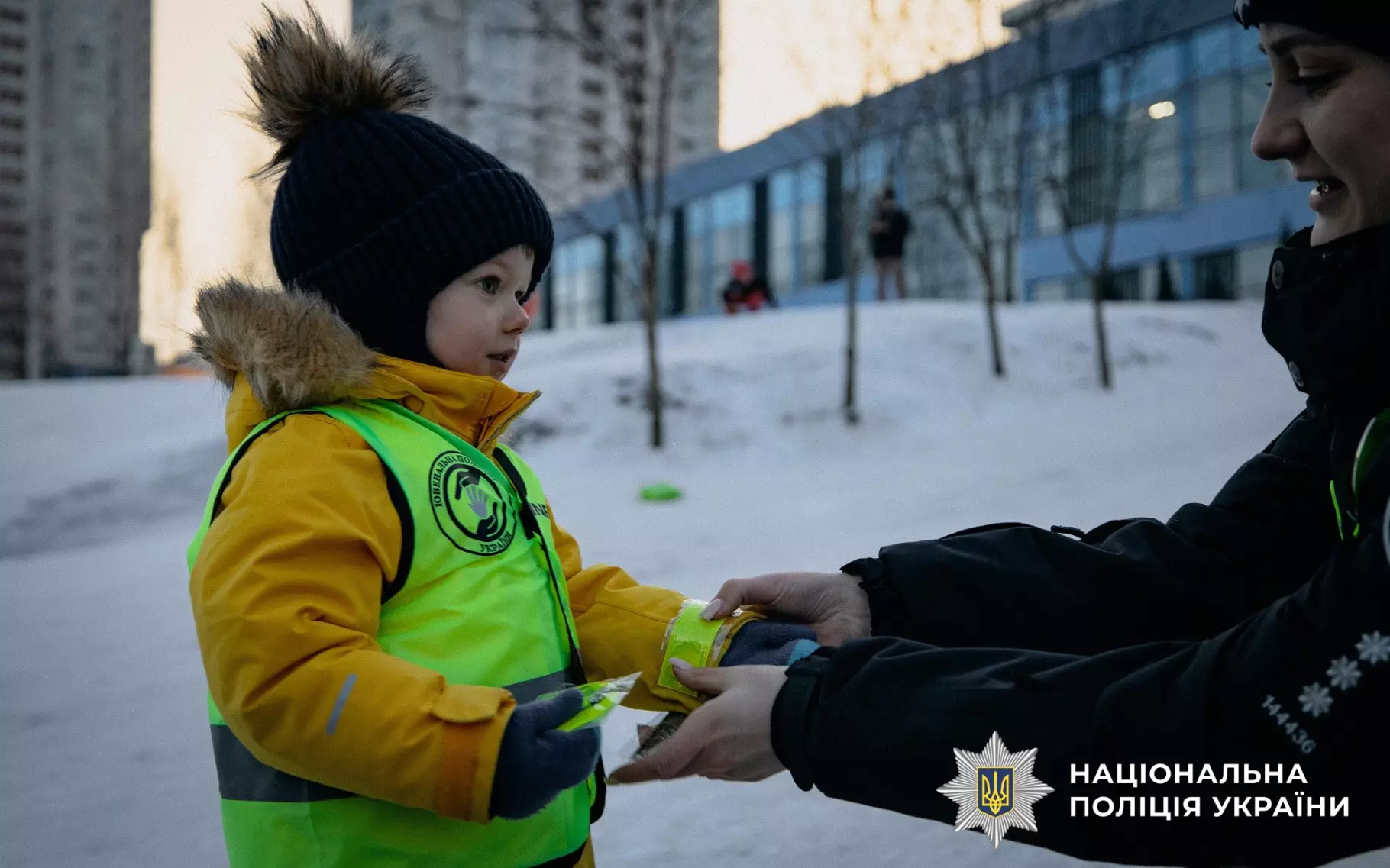 Дитина в жовтому жилеті отримує світловідбивальний елемент від поліцейського на зимовій вулиці