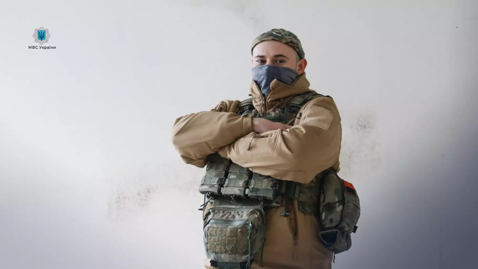 Прикордонник у військовій формі з маскою на обличчі стоїть зі схрещеними руками на тлі стіни