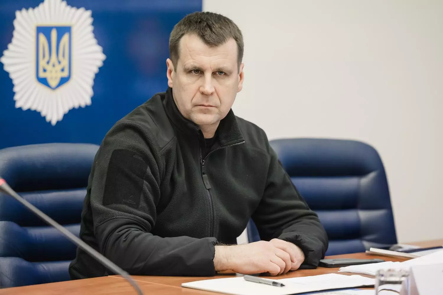 Чоловік у чорному одязі сидить за столом на офіційній зустрічі, на фоні герб України