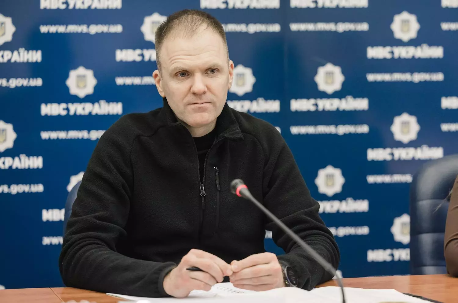 Чоловік у чорному светрі сидить за столом на офіційній зустрічі, на фоні логотипи МВС України