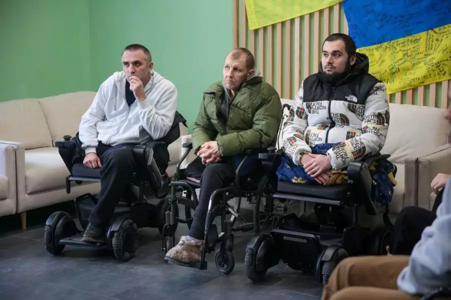 Троє поранених військових у візках сидять у кімнаті, на стіні прапор України з підписами