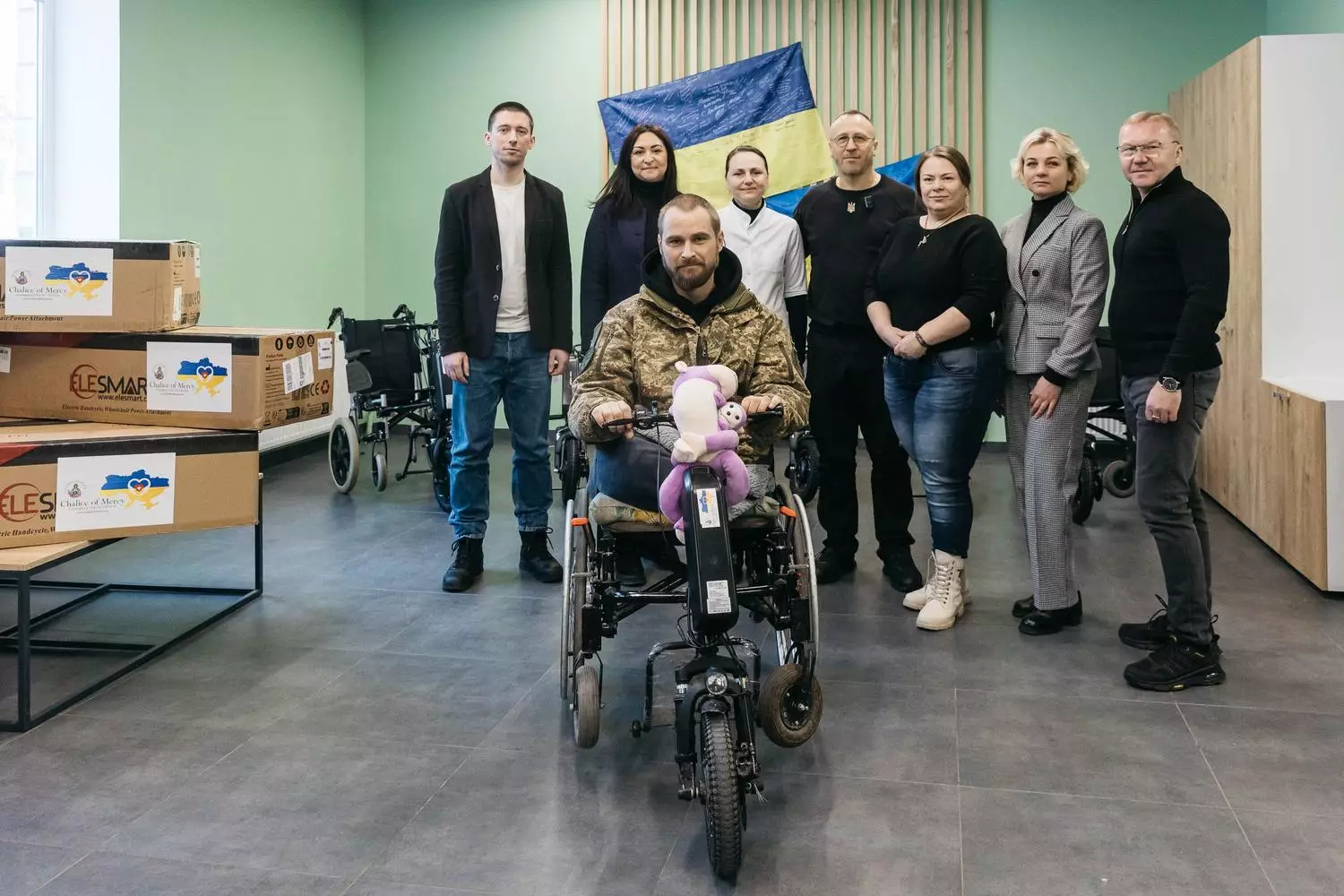Група людей з ветераном на електричному візку в приміщенні з українським прапором на стіні