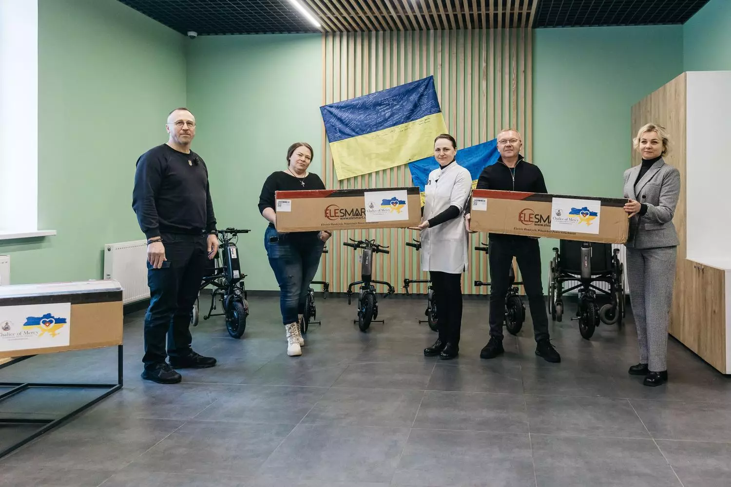 Група людей у приміщенні з коробками електроприводів для інвалідних візків на фоні прапора України