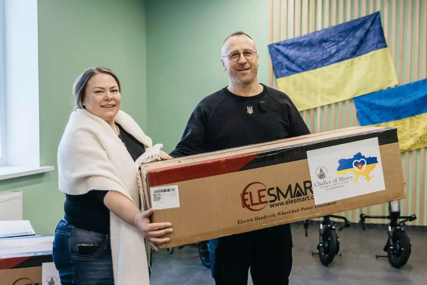 Чоловік і жінка тримають коробку з електроприводом для крісла колісного на тлі прапорів України