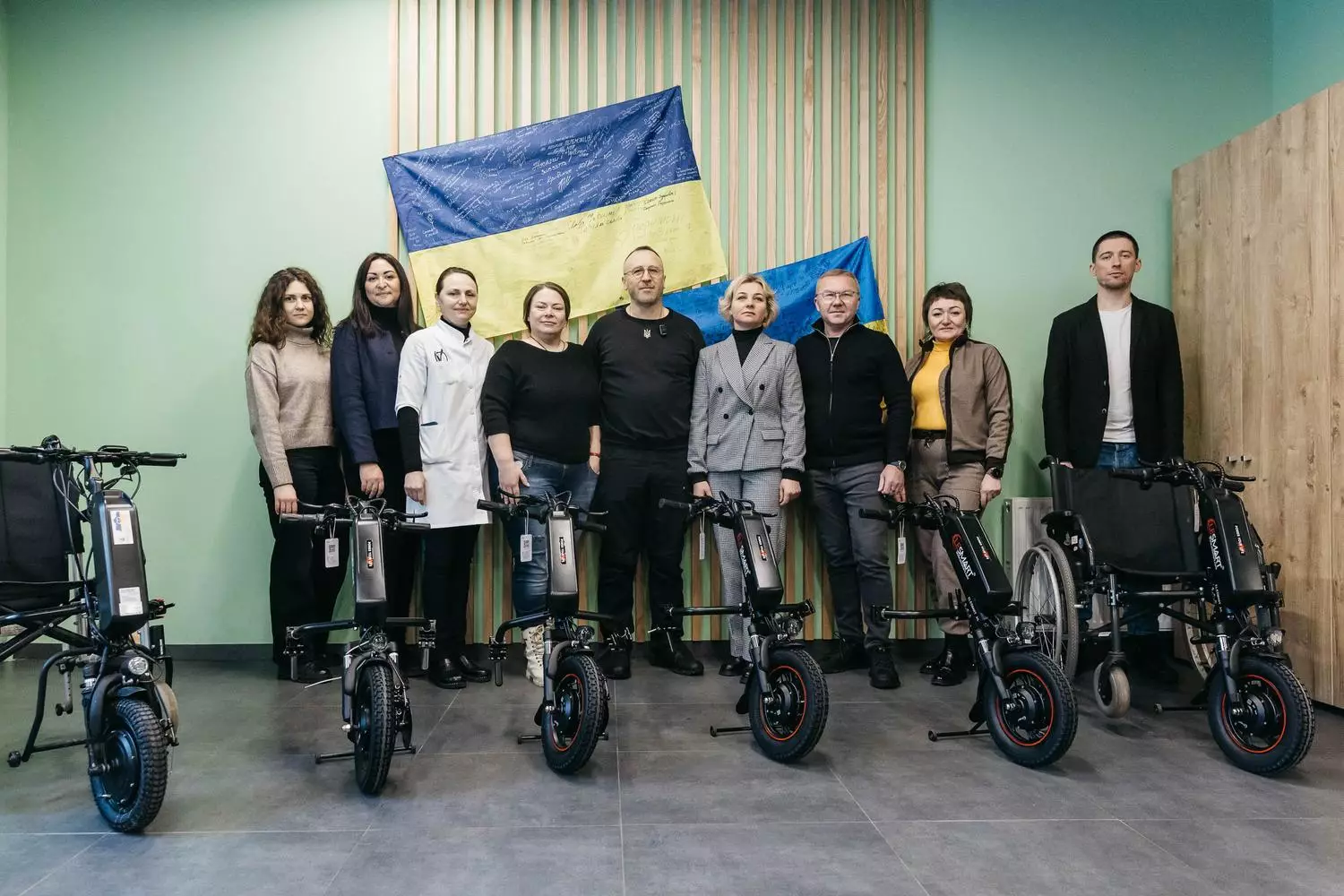 Група людей з електроприводами для крісел колісних на фоні українського прапора в медичному центрі