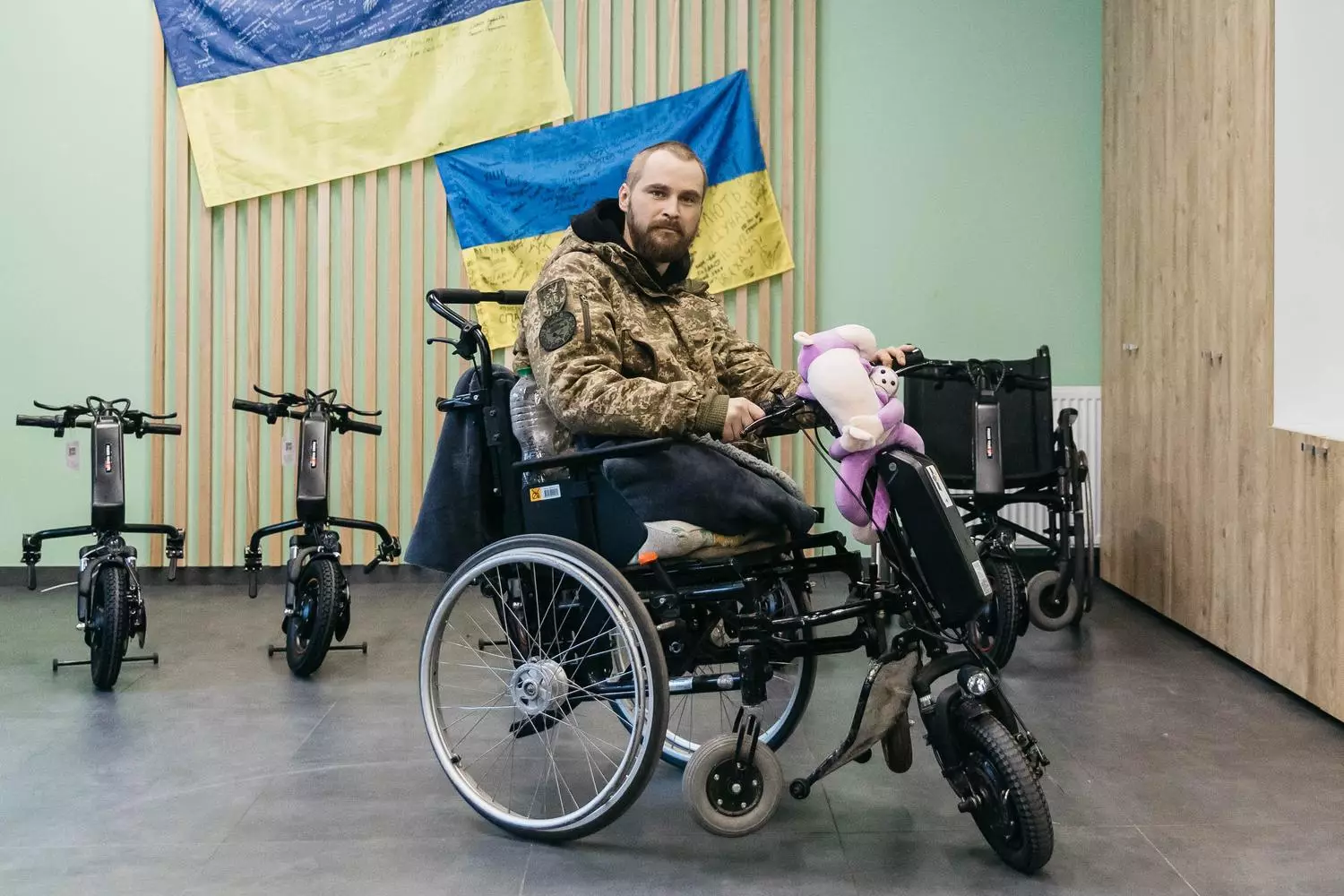 Чоловік у військовій формі на інвалідному візку з електроприводом, на тлі українських прапорів