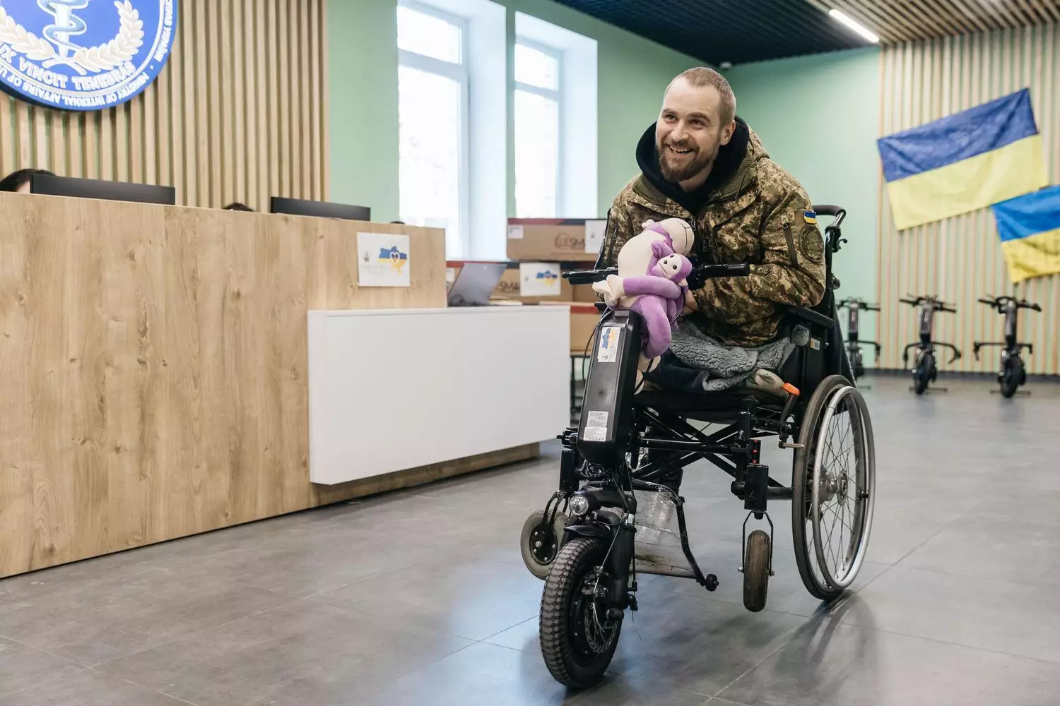 Чоловік у камуфляжі на інвалідному візку з електроприводом, усміхається в приміщенні з прапорами України