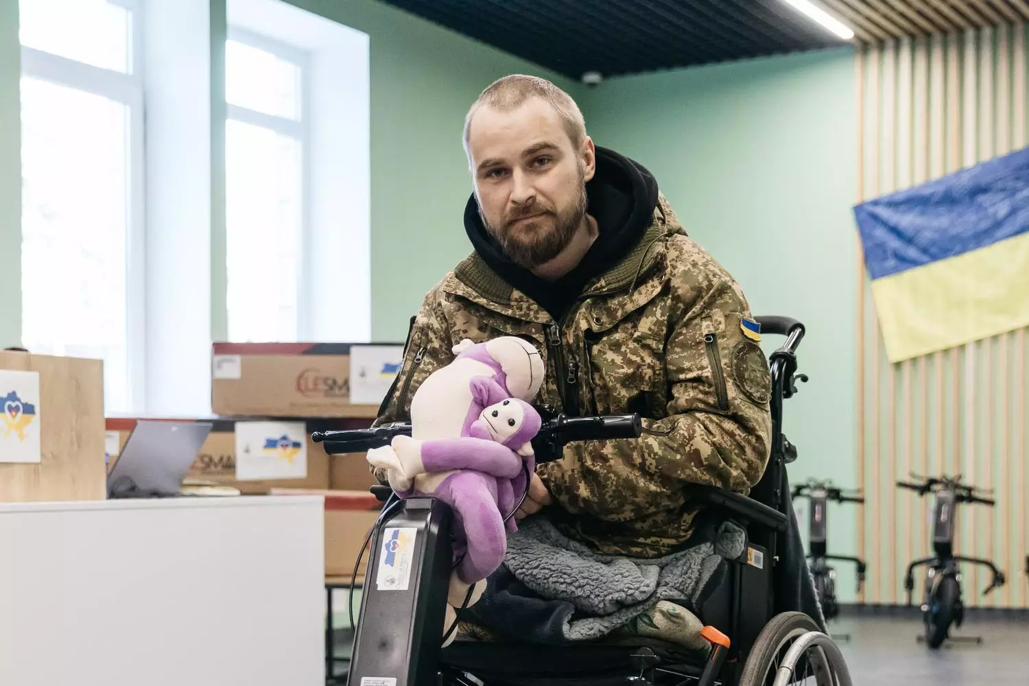 Чоловік у військовій формі сидить у кріслі колісному з електроприводом, тримаючи плюшеву іграшку