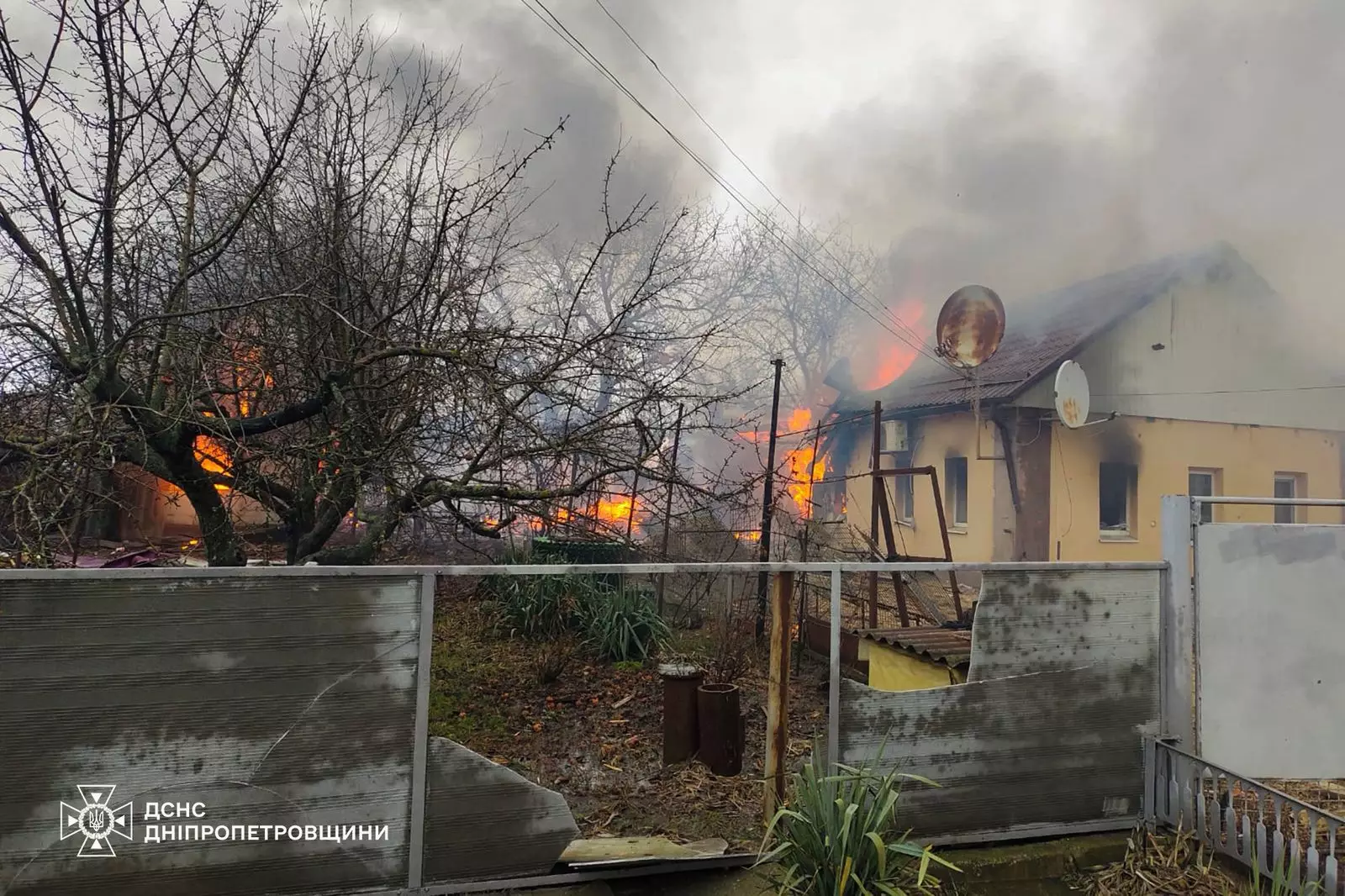 Палаючий будинок після обстрілу, густий дим піднімається вгору, пошкоджена огорожа на передньому плані