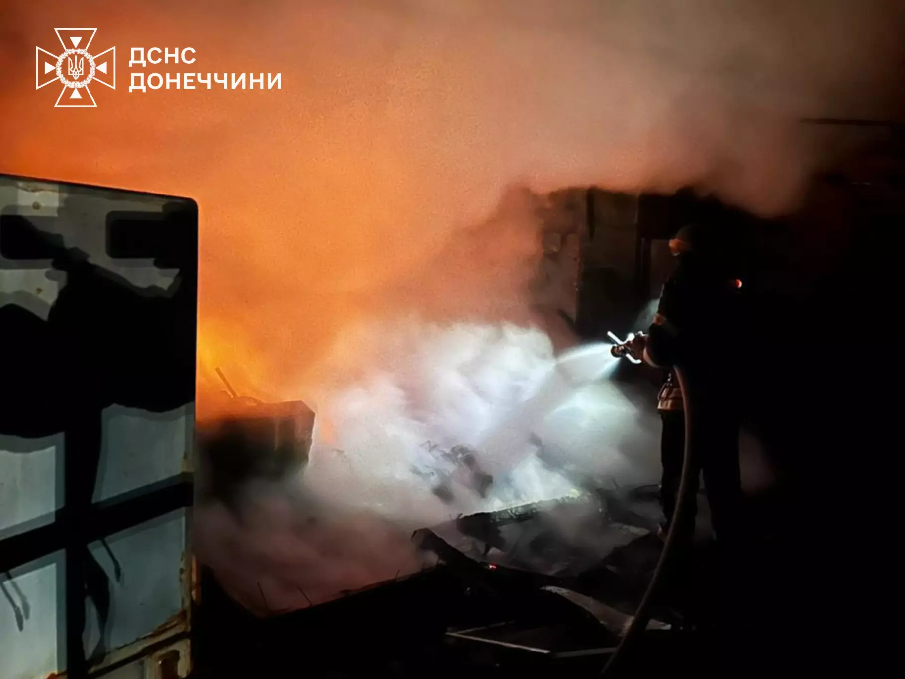 Рятувальник гасить пожежу вночі після обстрілу, густий дим і вогонь навколо, логотип ДСНС Донеччини