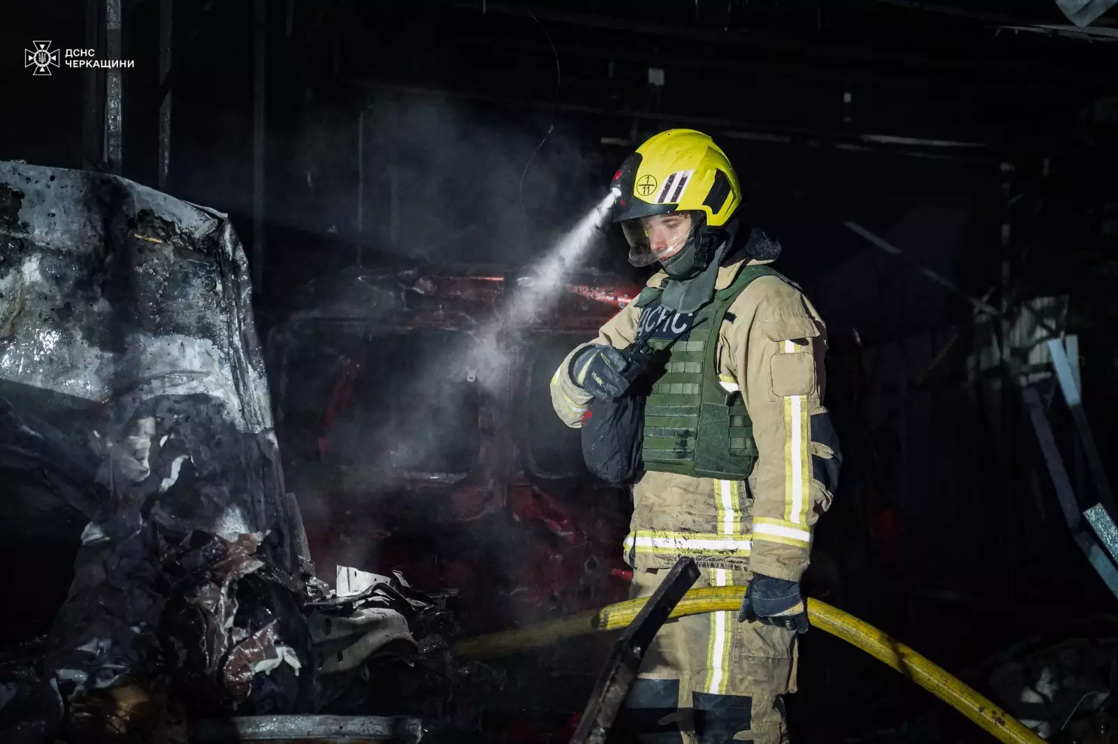 Рятувальник у захисному спорядженні гасить пожежу після обстрілу, темрява навколо, дим піднімається вгору