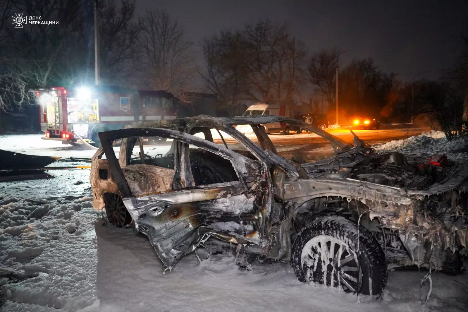Згорілий автомобіль на засніженій вулиці, пожежна машина на задньому плані, нічний час