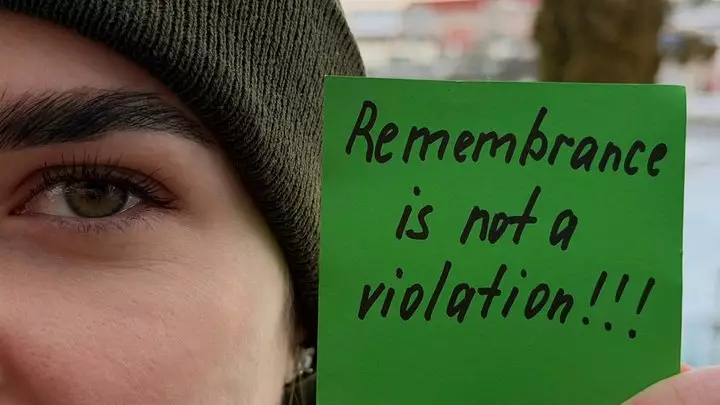 Людина в шапці тримає зелений папірець з написом "Remembrance is not a violation