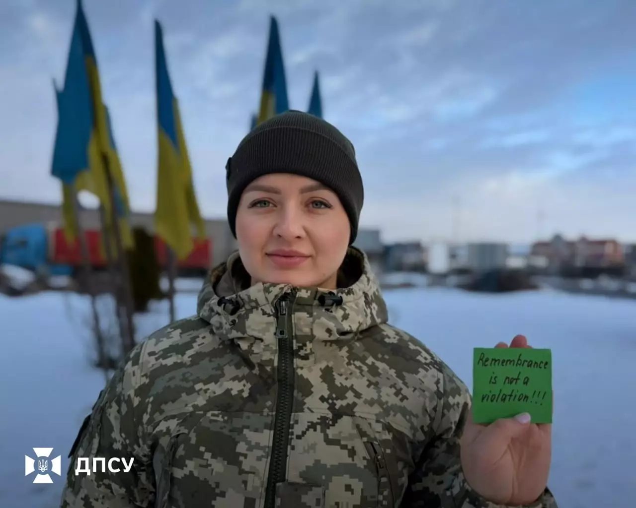 Прикордонник у камуфляжі тримає табличку з написом "Remembrance is not a violation" на фоні прапорів України