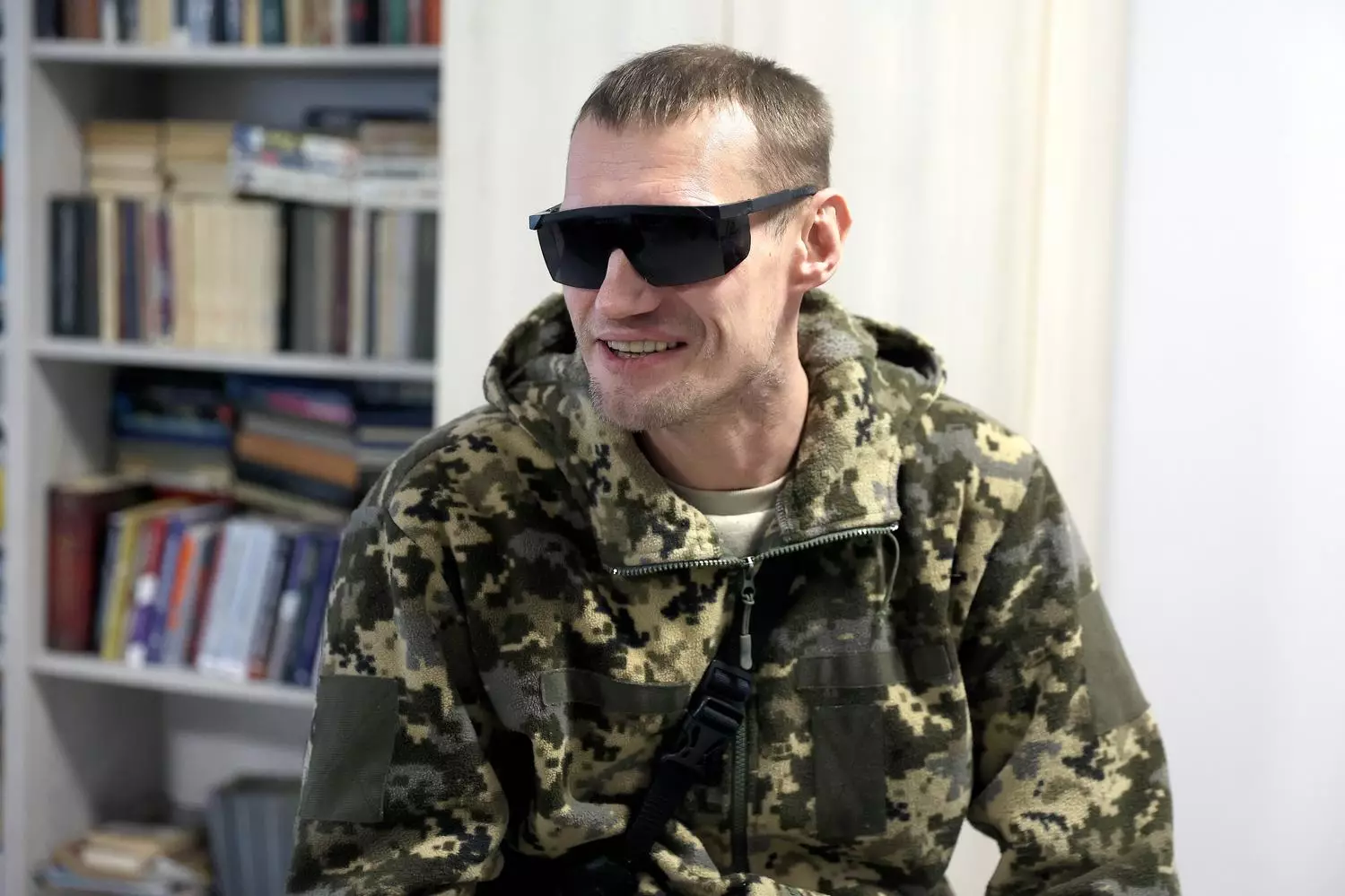 Ветеран у камуфляжі та темних окулярах сидить у кімнаті з книжковою шафою на задньому плані