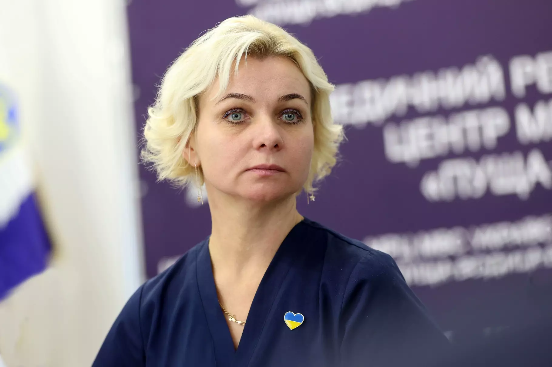 Жінка в темному одязі з жовто-блакитним значком на фоні банера медичного реабілітаційного центру