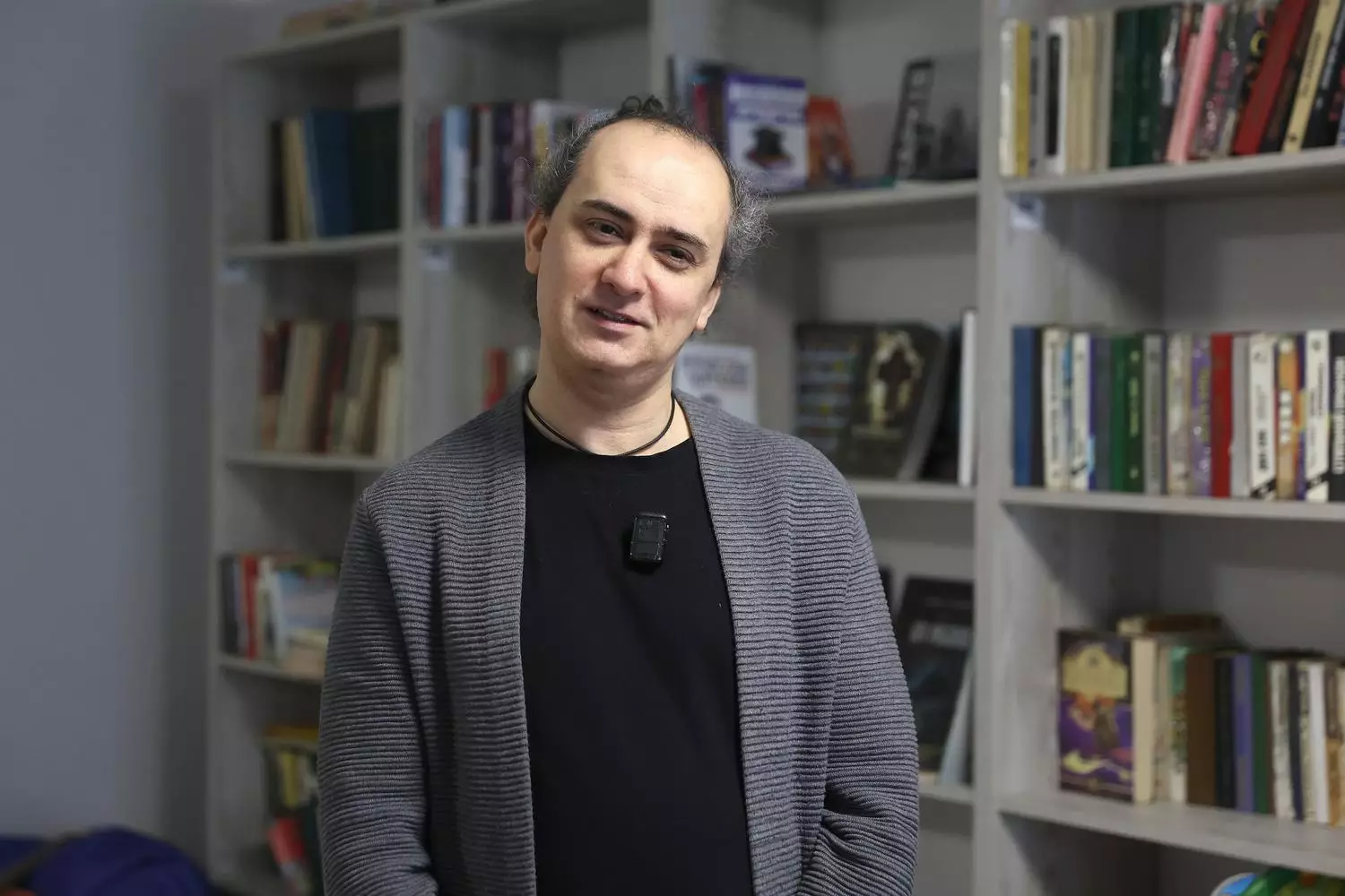 Чоловік у сірому кардигані стоїть у бібліотеці, оточений книжковими полицями, усміхається