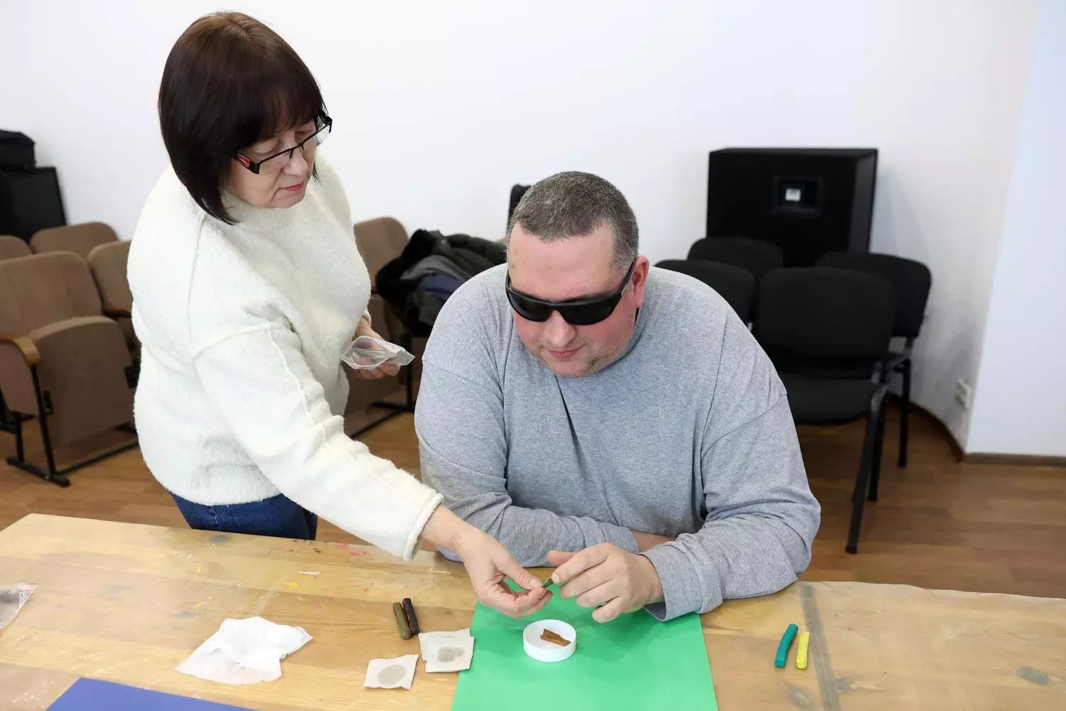 Чоловік у темних окулярах на занятті з арт-терапії в реабілітаційному центрі, жінка допомагає йому