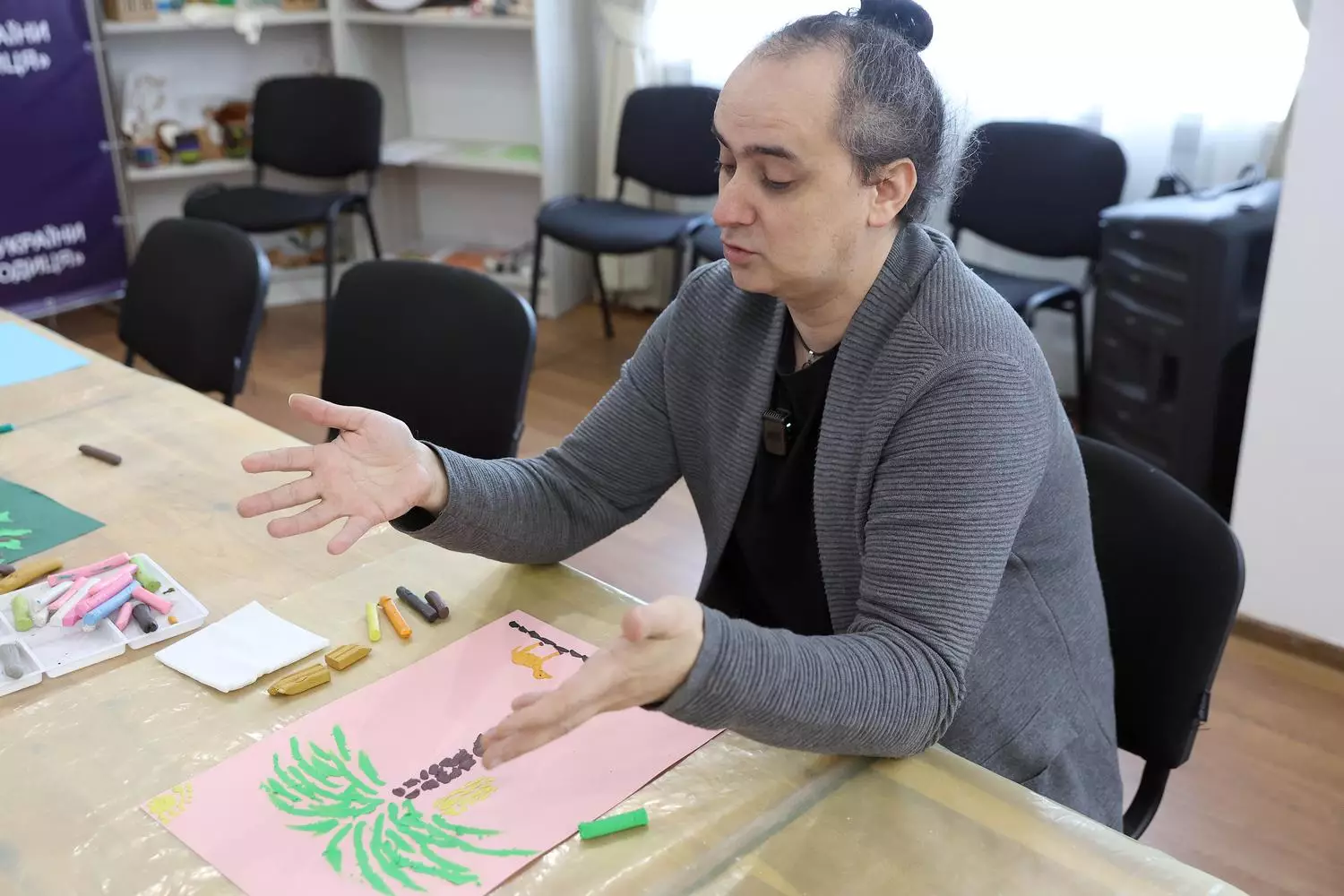 Чоловік у сірому светрі займається арт-терапією, малюючи пастеллю на рожевому аркуші в реабілітаційному центрі