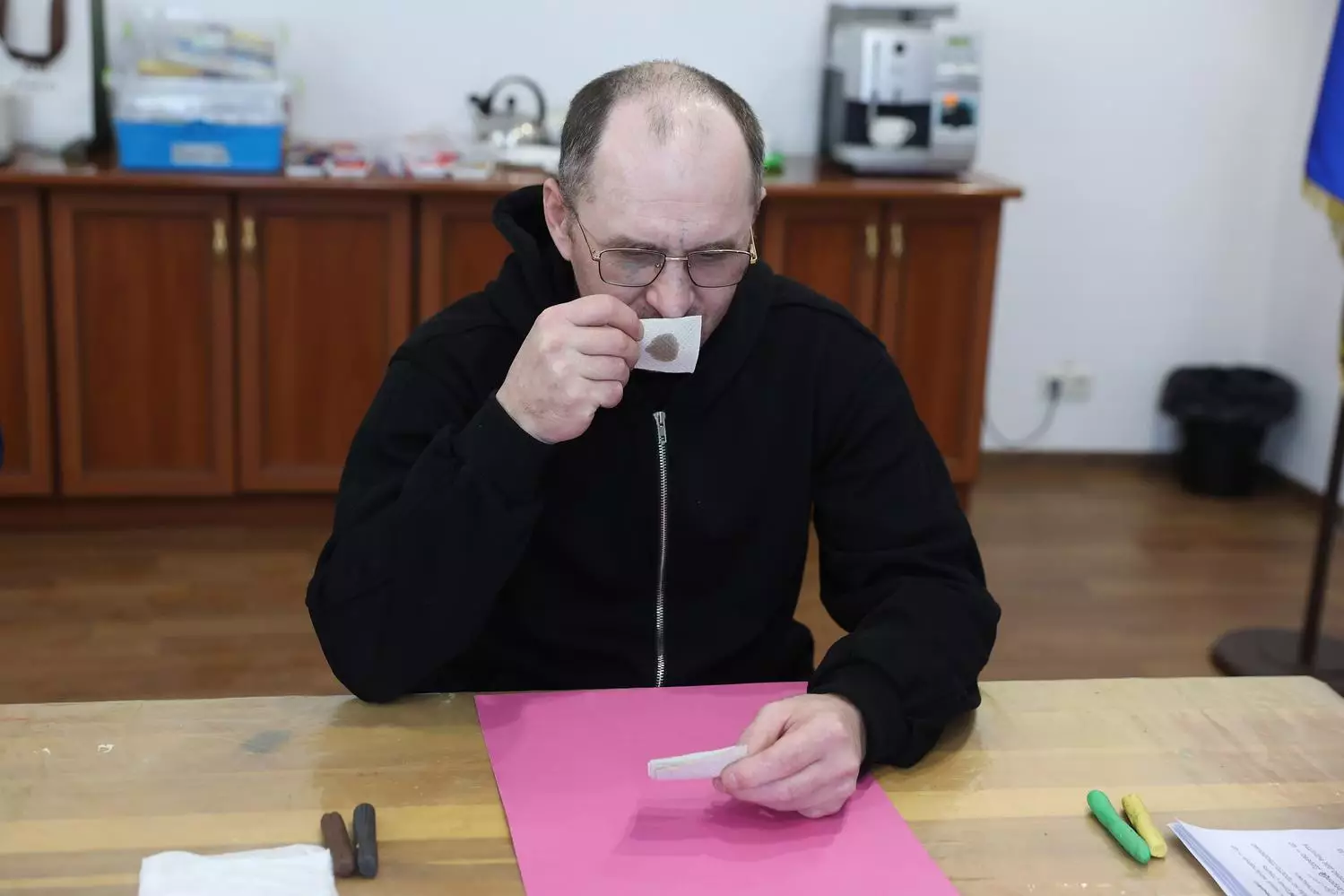 Чоловік у чорній кофті п'є чай за столом, тримаючи аркуш паперу, на фоні медичного центру