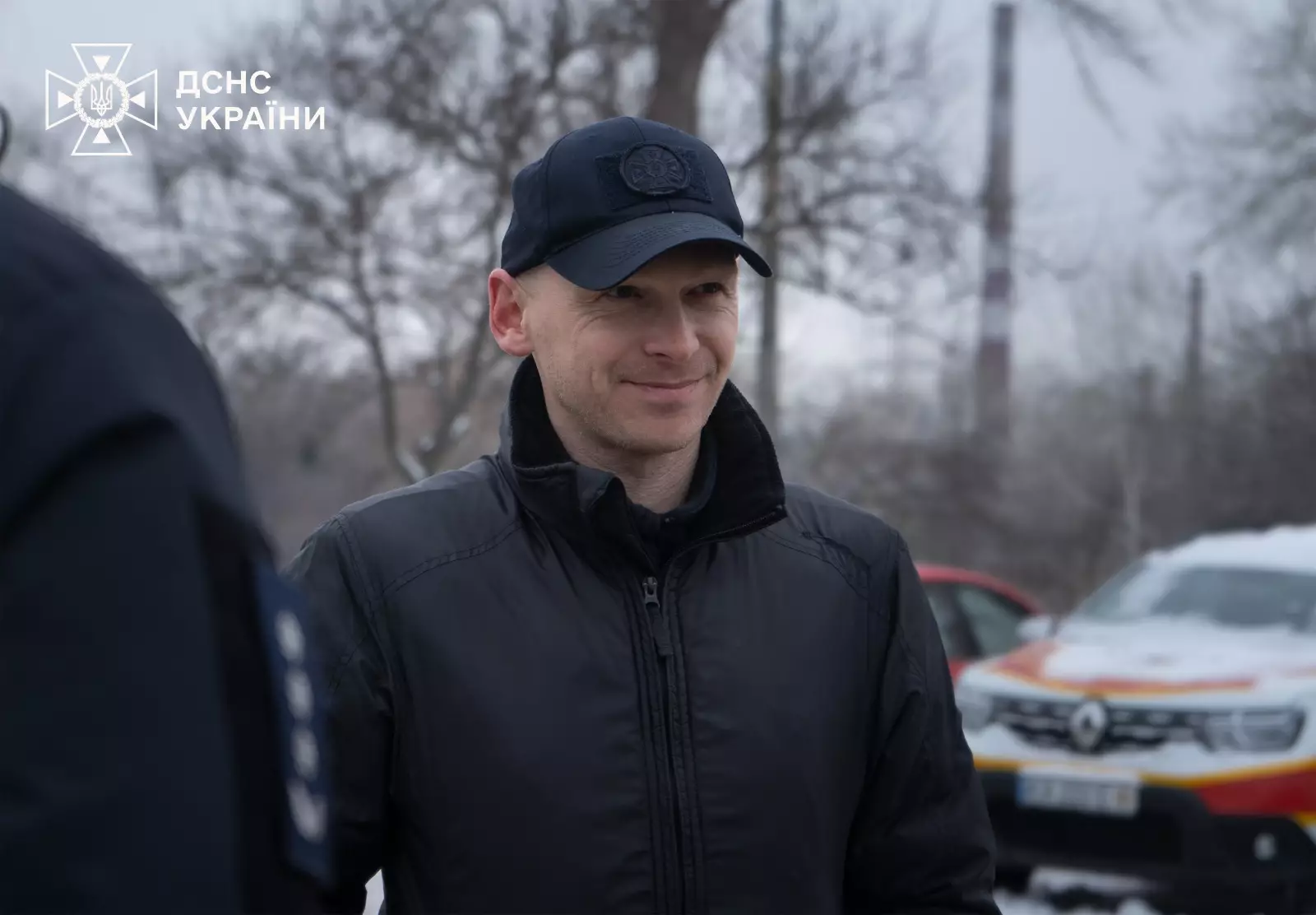 Чоловік у чорній куртці та кепці на фоні автомобіля ДСНС України під час церемонії передачі транспорту