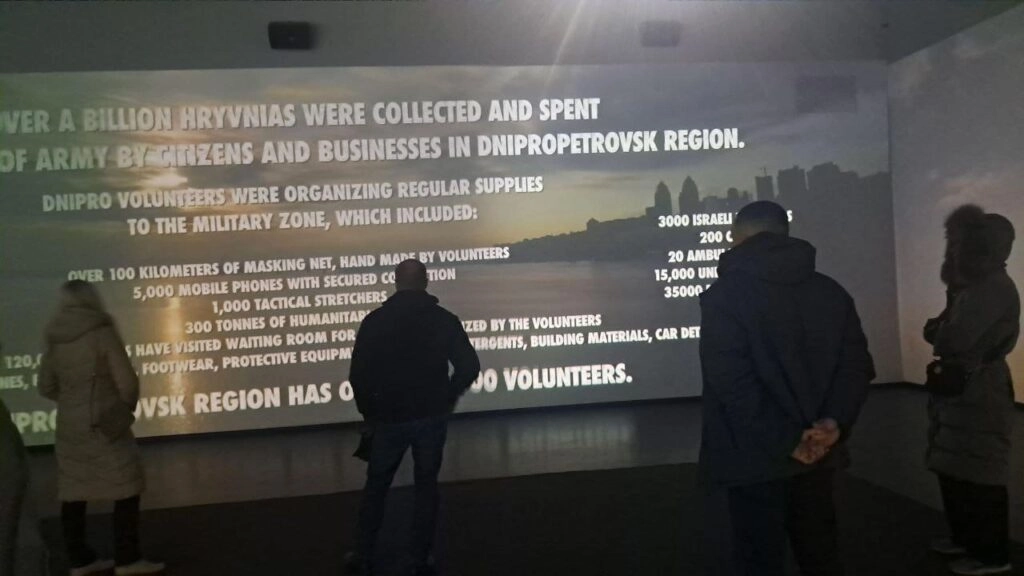 Люди стоять перед великим екраном з текстом про волонтерську діяльність у Дніпропетровському регіоні