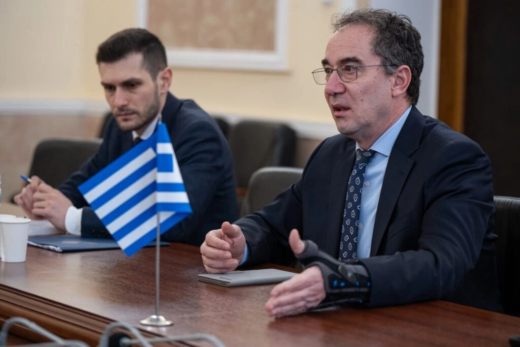 Двоє чоловіків за столом під час зустрічі, на передньому плані прапор Греції