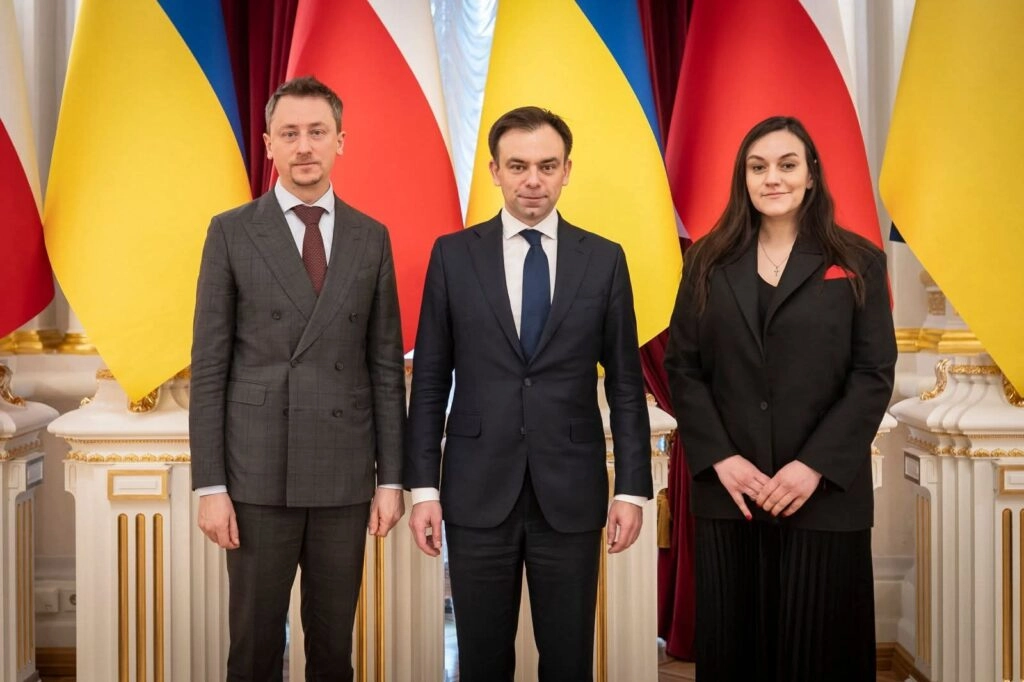 Троє людей у ділових костюмах стоять перед прапорами України та Польщі в офіційній залі