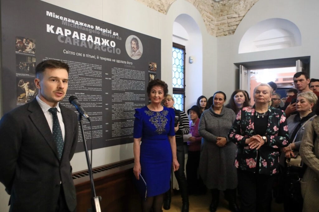 Група людей на виставці, присвяченій Караваджо, слухає промову перед інформаційним стендом